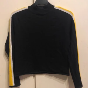 Svart tröja med gula och vita ränder från H&M - Säljer en svart tröja från H&M med coola gula och vita ränder längs ärmarna. Tröjan har en hög krage och är perfekt för en sportig look. Materialet är en blandning av viskos, akryl och polyamid vilket gör den både bekväm och hållbar.