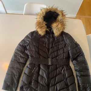 Dunjacka Vinterjacka Hollies 40 Dam - Använd en säsong, fint skick överlag, ett litet hål ca 2mm längst ned i fram till höger och något nött vid blixtlås-”flärp”.  Dun 80%, fjäder 20%. Fusk-päls i polyester vid huvan. Kemtvätt. Behöver en tvätt för att fräschas upp. 