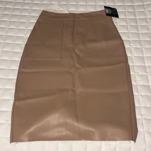   NLY Trend Nougat kjol -  Snygg nougatfärgad kjol i skinnimitation från NLY Trend. Kjolen har en stilren design med en slits baktill och stora fickor fram. Perfekt för både vardag och fest! Nyskick med prislapp kvar 🤎✨ 