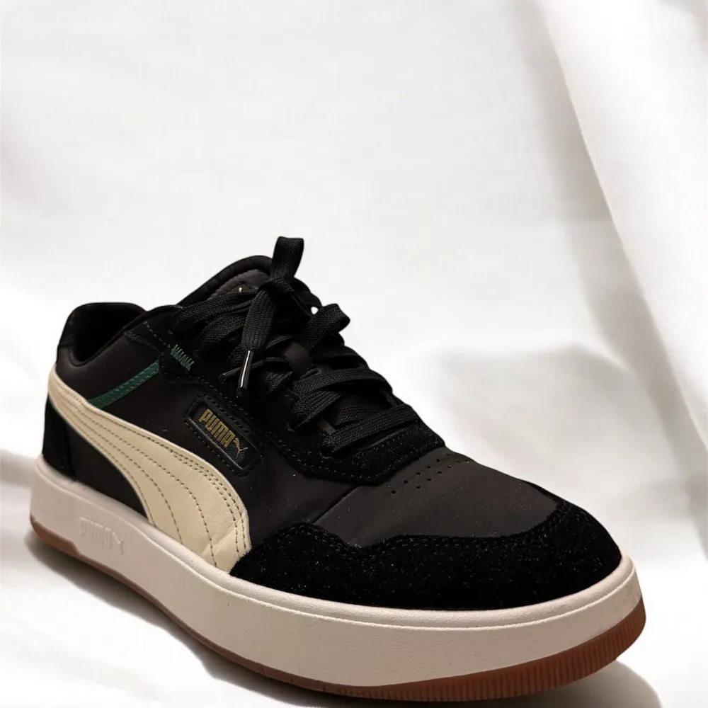 Snygga svarta sneakers från Puma i mocka och syntet. De har en krämfärgad sula och detaljer i beige och grönt. Perfekta för vardagsbruk med klassisk snörning och en stilren design.. Skor.