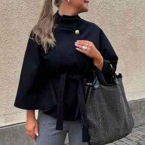 Svart kappa med knappar och bälte - Säljer en elegant svart poncho med hög krage och en snygg guldfärgad knappdetalj. Den har vida ärmar och ett bälte i midjan för en stilren look. Perfekt för höst och vår när du vill vara både varm och chic. Passar lika bra till vardags som till fest! Skärpet är tyvärr borta men man kan ha vilket som helst till. Helt oanvänd och nypris är 460kr. Onesize (ej mina bilder)