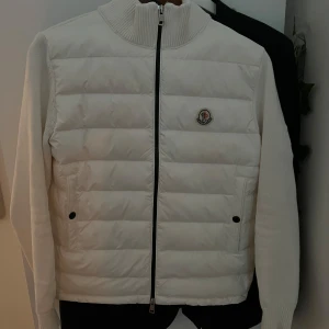 Vit moncler cardigan - Säljer en snygg vit cardigan från Moncler i mycket bra skick. Jackan har en quiltad design med dragkedja och ribbade muddar. Perfekt för kyliga höst- och vinterdagar. Den har långa ärmar och en stilren look med Moncler-logga på bröstet.