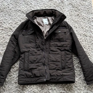 Brun jacka från J.Lindeberg - Säljer en snygg brun jacka från J.Lindeberg i storlek L. Jackan har en stilren design med dragkedja och knappar framtill. Den är perfekt för höst och vinter med sina långa ärmar och quiltade detaljer. Passar både till vardags och lite finare tillfällen.  Skickeg är i topp 9/10 inga defekter! 