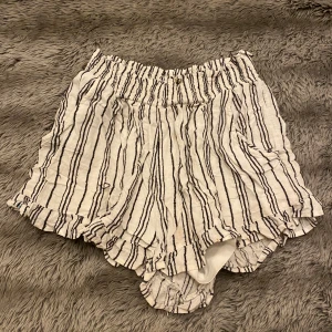 Randiga shorts med volangdetaljer - Superfina randiga shorts i vitt och svart med volangdetaljer. Perfekta för sommaren med en elastisk midja för extra komfort. De är gjorda i ett lätt och luftigt material, vilket gör dem idealiska för varma dagar. Passar både till stranden och en avslappnad dag på stan.