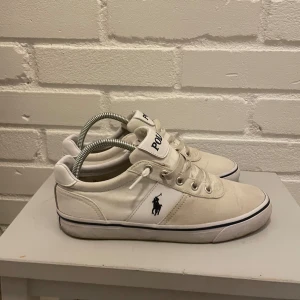 Vita sneakers från Polo Ralph Lauren - Säljer ett par vita sneakers från Polo Ralph Lauren i bra skick. De har en klassisk design med snörning och den ikoniska loggan på sidan. Perfekta för en avslappnad stil och passar till de flesta outfits.