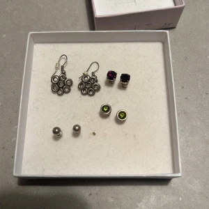 Set med olika örhängen - Säljer ett set med fyra olika par örhängen. Ett par är silverfärgade med hängande cirklar, ett par med mörkröda stenar, ett par med gröna stenar och ett par enkla silverkulor. Perfekt för att mixa och matcha till olika outfits!