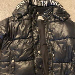 Svart pufferjacka från Calvin Klein Jeans - Säljer en snygg svart pufferjacka från Calvin Klein Jeans. Jackan har en hög krage med märkets logga och stängs med dragkedja och knappar. Perfekt för kalla dagar och ger en cool look. Passar både till vardags och fest!