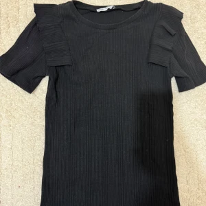 Svart ribbad topp från Zara - Snygg svart ribbad topp från Zara i storlek S. Den har korta ärmar med volangdetaljer.
