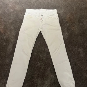 Vita byxor från Dsquared2 - Säljer ett par snygga vita byxor från Dsquared2. De har en klassisk design med knappar och dragkedja framtill. Byxorna är i bra skick och passar perfekt för både vardag och fest. Märkesdetaljer syns tydligt på både fram- och baksidan. Perfekta för dig som vill ha en stilren look!