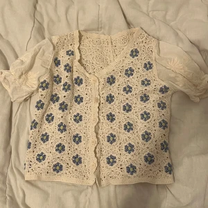 Beige virkad kofta med blommönster - Superfin beige virkad kofta med blå blommor. Den har korta ärmar och en söt, vågig kant framtill. Perfekt för vår och ger en romantisk touch till vilken outfit som helst. Passar både till vardagar 