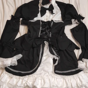 Svart och vit Lolita Klänning med spetsdetaljer - Säljer en unik svart och vit klänning med spetsdetaljer och snörning framtill. Klänningen har långa ärmar och volanger, perfekt för en speciell tillställning. Den har en klassisk look med en modern twist. Passar perfekt för höst och vinter. Det är en Lolita inspirerad klänning En lite större S eller mindre M. Armarna kan man plocka bort.