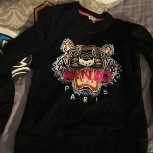 Svart tröja med tigerbroderi från Kenzo - Säljer en svart tröja från Kenzo i storlek XS. Den har ett coolt tigerbroderi på framsidan med texten 'Kenzo Paris' i rosa och vita bokstäver. Perfekt för att ge din outfit en edgy touch! 🐯