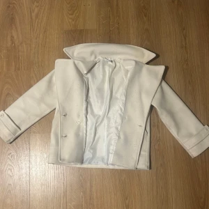 Beige kappa - Säljer en stilren krämvit kappa i mycket bra skick. Den har en klassisk design med dubbelknäppning och långa ärmar. Perfekt för höst och vår, och funkar lika bra till vardags som till fest. Nypris 757kr (Design By Si). 