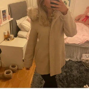 Beige kappa med fuskpäls - Säljer en superfin beige kappa med fuskpäls på huvan. Den är perfekt för höst och vinter och har en mysig känsla. Kappan är långärmad och har en stilren design som passar till det mesta. Använd fåtal gånger så mycket bra skick, köpt för 800kr🥰