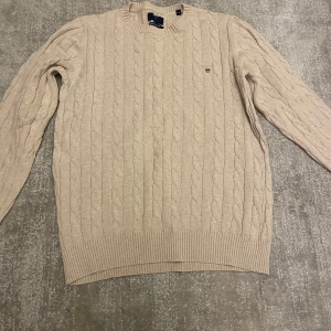Beige stickad tröja - Säljer en snygg beige GANT tröja med kabelstickat mönster. Den har långa ärmar och ribbade muddar vid ärmslut och nederkant. Perfekt för kyliga dagar! bara att höra av sig vid intresse!