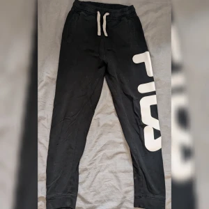 Svarta mjukisbyxor från Fila - Säljer ett par svarta mjukisbyxor från Fila. De har en bekväm passform med snörning i midjan och ett stort vitt Fila-tryck på benet. Perfekta för en avslappnad dag hemma eller på gymmet. 