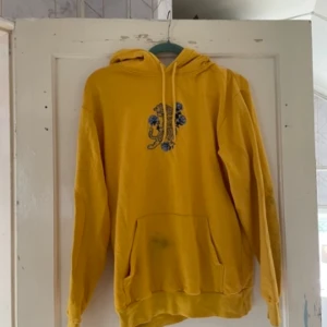 Gul hoodie från Divided - Säljer en superfin gul hoodie från Divided. Den har en cool blå blommig detalj på bröstet och en klassisk känguruficka framtill. Perfekt för en avslappnad stil och passar bra till både jeans och leggings. Den har dock fläck på fickan går kanske bort
