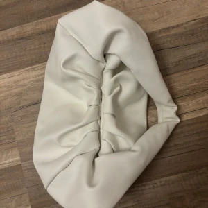 Vit axelväska i syntetmaterial - Säljer en stilren vit axelväska i syntetmaterial. Väskan har en unik design med en vriden form och en mjuk struktur. Insidan är brun och har en dragkedja för säker förvaring. Perfekt för både vardag och fest!