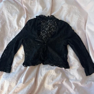 Svart spetsblus med volangdetaljer - Säljer en svart spets bolero/kofta med trekvartsärmar och fina volangdetaljer. Blusen har en knytning framtill och är perfekt för festliga tillfällen.🤩