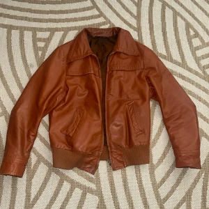 Brun skinnjacka med dragkedja - Säljer en snygg vintage brun skinnjacka. Den har ribbande muddar vid ärmslut och nederkant för en skön passform. Perfekt för höst och vår, och funkar lika bra till vardags som till fest. Jag brukar ha storlek M-L, så den kan se lite små ut på mig.