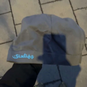 Grå mössa med broderad text - Säljer en grå mössa med en cool broderad text i ljusblått. Perfekt för att hålla sig varm under kyliga dagar. Den är mjuk och bekväm, perfekt för vardagsbruk.