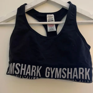 Svart sporttopp från Gymshark - Säljer en svart sporttopp från Gymshark i storlek M. Toppen är ärmlös och har en bred resår med Gymshark-loggan i vitt runtom. Perfekt för träning och ger bra stöd. Använd men i bra skick!