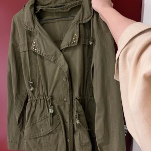 Olivgrön parkajacka med nitar - Olivgrön parkajacka med nitar på kragen. Jackan har en justerbar midja med dragsko och praktiska fickor framtill. Perfekt för höst och vår! Den har långa ärmar och en liten amerikansk flagga som detalj på ärmen. Luvan går att fälla in. Köpt i Candem 