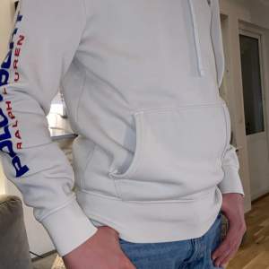 Säljer en snygg vit hoodie från Ralph Lauren med 'Polo Sport' tryckt i blått och rött längs ärmen. Den har en klassisk känguruficka. Perfekt för en casual look eller träning!