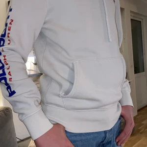 Vit hoodie från Ralph Lauren - Säljer en snygg vit hoodie från Ralph Lauren med 'Polo Sport' tryckt i blått och rött längs ärmen. Den har en klassisk känguruficka. Perfekt för en casual look eller träning!