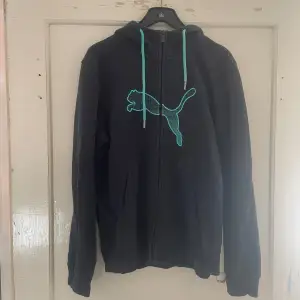 Säljer en svart hoodie från Puma i storlek S. Den har en dragkedja framtill och en stor turkos Puma-logga på bröstet. Huvan har turkosa dragsnören som matchar loggan. Perfekt för en avslappnad stil eller träning.