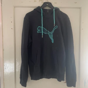 Svart hoodie från Puma - Säljer en svart hoodie från Puma i storlek S. Den har en dragkedja framtill och en stor turkos Puma-logga på bröstet. Huvan har turkosa dragsnören som matchar loggan. Perfekt för en avslappnad stil eller träning.