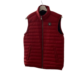 Röd quiltad dunväst - Säljer en snygg röd quiltad dunväst med dragkedja framtill. Västen har en hög krage och två fickor på sidorna. Perfekt för kyliga höst- och vårdagar. Den är i nyskick och passar bra för både vardag och friluftsliv.