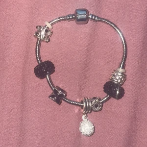 Charmigt armband med svarta och silver detaljer - Säljer ett superfint armband med en mix av svarta och silverfärgade berlocker. Armbandet har flera olika charms, glittrig boll som hänger ner, en blomformad charm och flera med mönstrade detaljer. Man kan ta av och på de berlocker man inte gillar!