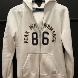 Vit hoodie från Peak Performance - Säljer en snygg vit hoodie från Peak Performance i storlek M. Den har en dragkedja framtill och en cool '86' och 'Peak Performance' text på bröstet. Perfekt för en avslappnad stil med ribbade muddar och en skön huva med ett 'P' på baksidan. Grym för både vardag och chill!