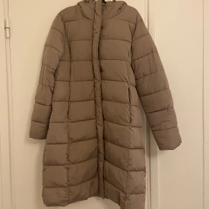 Vinter jacka  - Säljer en vinterjacka från Lager 157 i storlek L, använd endast förra vintern. Jackan är beige/khaki och lång, perfekt för vintern. Den har både dragkedja och knappar för extra skydd, samt en luva som håller dig varm. Jackan är i mycket bra skick! 😊