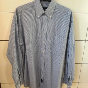 Blå och vit rutig skjorta från GANT - Säljer en klassisk blå och vit rutig skjorta från GANT i storlek M. Skjortan är tillverkad i 100% bomull och har långa ärmar med knappar. Perfekt för både vardag och festliga tillfällen!