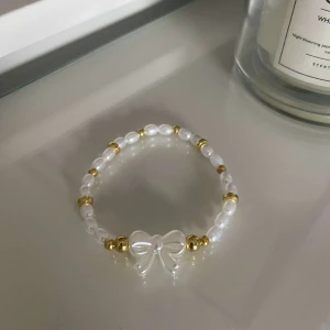 Vitt pärlarmband med rosett - Säljer ett supergulligt armband med vita pärlor och gulddetaljer. Armbandet har en söt rosett i mitten som ger det en extra charmig touch. Perfekt för att piffa upp vilken outfit som helst!