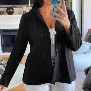 Svart jacka med huva och dekorativa knappar - stilren svart A formad jacka från Zara . Den har en praktisk huva och dekorativa knappar framtill som ger en unik touch. Jackan är perfekt för både vardag och lite finare tillfällen. Den är i bra skick. 