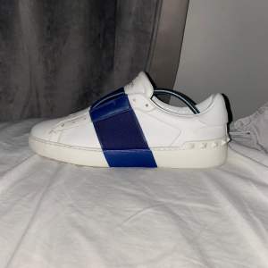 Säljer ett par snygga Valentino sneakers i vitt med en cool blå rem över ovansidan. Skorna är i skinn och har en stilren design med diskreta nitar på sidorna. Perfekta för både vardag och fest!