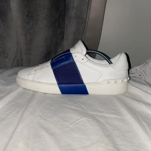 Valentino sneakers med blå detalj - Säljer ett par snygga Valentino sneakers i vitt med en cool blå rem över ovansidan. Skorna är i skinn och har en stilren design med diskreta nitar på sidorna. Perfekta för både vardag och fest!