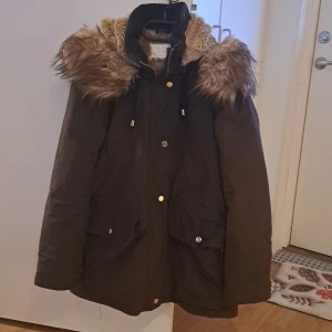 Grön parkajacka med fuskpäls - Säljer en snygg grõn parkajacka med fuskpäls på huvan. Jackan har en varm och mysig foder inuti, perfekt för kalla vinterdagar. Den har långa ärmar och praktiska fickor med både dragkedja och knappar. Kedjan är sönder men annars det funkar perfekt 👌