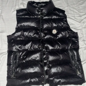Svart dunväst från Moncler - Säljer en snygg svart dunväst från Moncler. Västen har en glansig finish och är quiltad för extra värme. Den har en dragkedja framtill och två praktiska fickor med dragkedjor. Perfekt för kyliga höst- och vinterdagar!