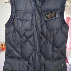 Babour marinblå väst - Marinblå väst med fickor framtill och små fickor på bröstet. Västen har en logga på vänster bröst. Längd 65 cm. Använd ett fåtal gånger.