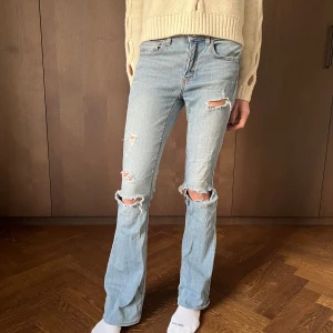 Lowwaist bootcut jeans - Säljer ett par ljusa jeans med slitningar på knäna och låren. De har en bootcut-stil och är lowwaist, vilket ger en snygg siluett. Perfekta för en avslappnad look med en stickad tröja eller t-shirt. Passar bra till både vår och höst!