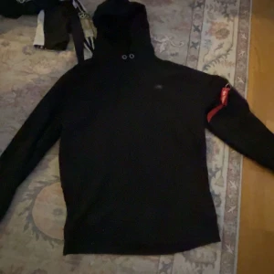 SvartAlhpa hoodie med ficka på arm. - Säljer en svart hoodie med en cool röd 'Alpha detalj.Liten ficka med dragkedja och är perfekt för en avslappnad stil. Passar bra till både vardag och chillkvällar. Hör av dig om du är intresserad! 🖤