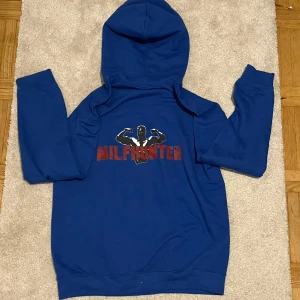 Blå MILFHUNTER hoodie. - Säljer en blå MILFHUNTER hoodie från Revenite i storlek M. Den har en cool design med texten ”MILFHUNTER” och den säljs inte längre! Perfekt för en avslappnad stil eller som statement-plagg. Gjord i ett mjukt och bekvämt material.