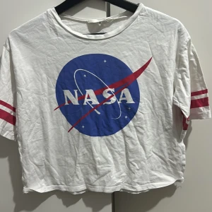 Vit NASA t-shirt från H&M - Säljer en cool vit t-shirt från H&M med NASA-logga i blått och rött. Den är croppad och har korta ärmar med röda ränder. Perfekt för en avslappnad stil eller för att visa din kärlek till rymden! 🚀