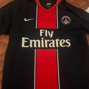 Säljer en snygg svart PSG fotbollströja från Nike med röd och blå detalj i mitten. Tröjan har korta ärmar och Fly Emirates som sponsorlogga på framsidan. Perfekt för alla PSG-fans! ⚽