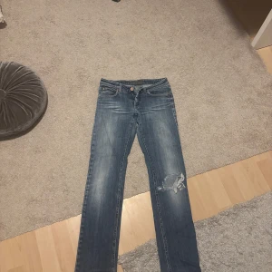 Blå jeans med slitningar - Säljer ett par snygga blå jeans med slitningar på ena benet. De har en klassisk femficksdesign och en knappgylf. Perfekta för en avslappnad vardagslook!