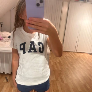 Vit t-shirt från GAP - Säljer en vit t-shirt från GAP i bomull. Den har en klassisk passform med korta ärmar och GAP-loggan tryckt i svart på framsidan. Perfekt för en avslappnad stil eller sommarhäng. Skön och enkel att matcha med jeans eller shorts!